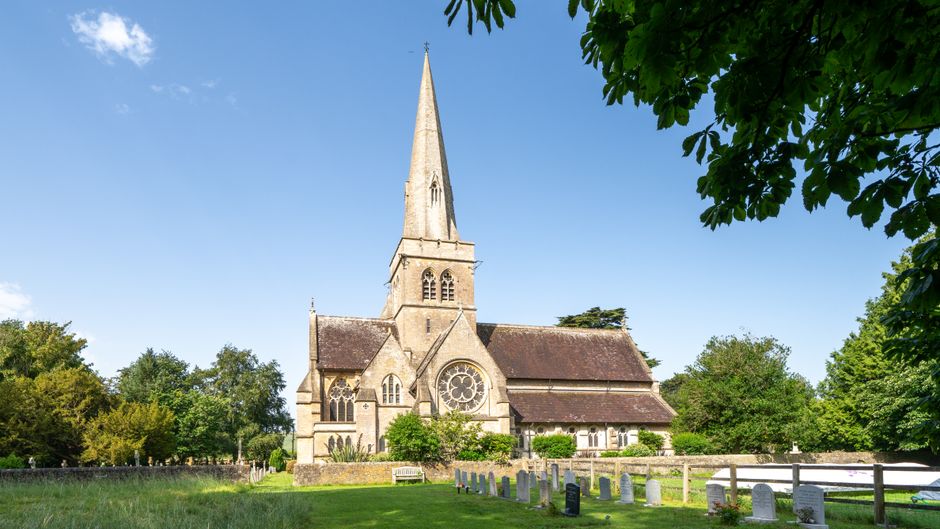 A historic stone church with a tall, pointed spire and Gothic architectural features, including arched windows and intricate tracery, is situated within a green surrounding that includes trees with leafy branches and a grass lawn. In the foreground, a small graveyard with evenly spaced white and grey headstones is visible, enclosed by a wooden fence. The church is bathed in natural daylight against a clear blue sky, with sunlight highlighting the textured stone façade and foliage, creating a serene, well-maintained environment. This setting exemplifies a typical rural or village churchscape, often requiring private or independent waste clearance services to manage maintenance and grounds upkeep, which companies like Waste Clearance Sutton might assist with for local community sites.