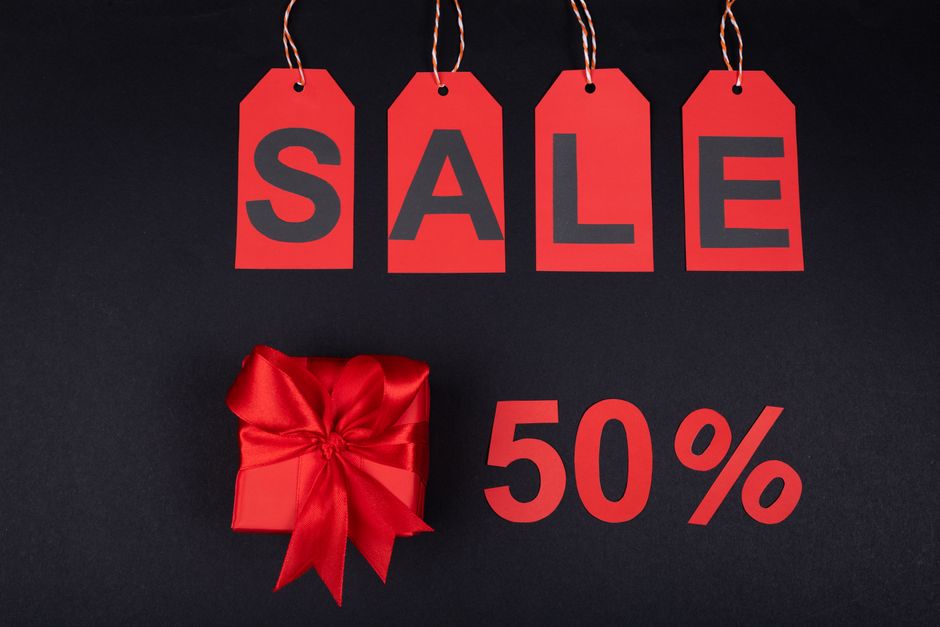 Four red paper tags, each with a black letter spelling the word 'SALE,' hang in a row against a dark background, attached to red and white striped string. Below the tags, a small, neatly wrapped red gift box with a shiny satin ribbon bow sits on the surface, accompanied by bold red text indicating '50%.' The scene is well-lit, highlighting the vibrant red tones of the sale tags, gift box, and text, creating a visually striking promotional display that could relate to retail pricing or special offers. The overall composition emphasizes the theme of discount campaigns, which may be associated with private sales or clearance events, connecting subtly to the concept of alternative waste handling by illustrating a promotional context, although the primary focus remains on the sale display. The scene is clean, simple, and professional, with a focus on the visual impact of the sale elements while maintaining an informative tone suitable for an accessible description.