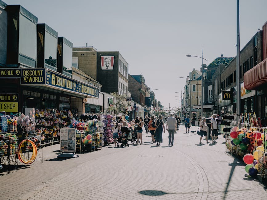 A busy pedestrian street scene in daytime with storefronts and outdoor stalls on both sides, including a discount shop and casual retail outlets. The shops display colorful merchandise, such as toys, balloons, and fabric goods, arranged in baskets and on racks. Several people are walking along the paved street, some pushing strollers or carrying shopping bags, while others browse the displays. The background features a mix of multi-storey buildings with signage, including familiar fast-food outlets and retail signs, set under a clear, bright sky. Streetlights line the walkway, and the environment suggests a lively urban shopping area, possibly undergoing an informal clearance or de-cluttering, typical of community markets or outdoor markets. The scene is well-lit with natural sunlight, casting shadows on the pavement, emphasizing the activity typical of a busy high street or marketplace that may relate to additional services like rubbish removal for clearing clutter or waste generated by retail and market activities, naturally aligning with the themes of private waste handling and on-site clearance solutions offered by Waste Clearance Sutton.