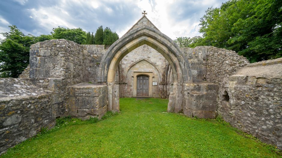 An ancient stone building entrance featuring a pointed Gothic archway, constructed from weathered grey stone blocks with varying textures and some visible mortar joints. The arch is framed by a series of stepped stone moldings, and the entrance is flanked by low, uneven stone walls extending to the sides, which appear partially ruined or aged. In front of the doorway, there is a small, well-maintained patch of green grass, contrasting with the rough stonework. Behind the structure, lush green trees and foliage are visible under a partly cloudy sky, with patches of sunlight illuminating the scene. The environment suggests an outdoor setting, possibly a historical site or ruins, with the surrounding natural landscape providing a peaceful atmosphere. The composition emphasizes the historic architecture and the interplay of natural elements, fitting within the context of a property that might require external maintenance or clearance services, such as waste clearance, described on wasteclearancesutton.co.uk.