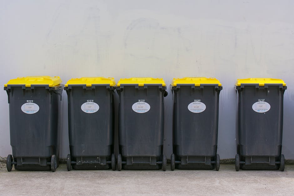 Five large wheeled rubbish bins with black bodies and yellow lids are lined up against a plain, light-colored wall. Each bin has a white oval label with a barcode and text, positioned towards the upper front section. The bins are situated on a concrete surface, with the first two on the left slightly closer together and the remaining three placed with some space between them. The environment appears to be a managed outdoor area, possibly a designated storage spot for waste collection. The bins are intended for general waste disposal, and their sturdy plastic material with a textured finish suggests they are designed for frequent use. Waste Clearance Sutton’s service focus on efficient rubbish collection is subtly supported by this scene, highlighting the typical equipment involved in private waste handling and on-site clearance activities. The lighting is neutral and even, providing clear visibility of each bin’s details, emphasizing the practicality and organization of waste storage for disposal or collection purposes.