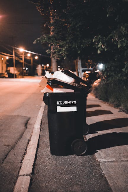 A black wheelie bin with a white label displaying 'ST. JOHN'S' is positioned on a pavement beside a residential street at night. The bin is filled with various types of waste, including cardboard boxes and plastic items, some of which are partially visible at the top and leaning over the bin's edges. The street is illuminated by warm, yellow streetlights, casting shadows on the pavement and nearby parked cars. In the background, there are trees with dense foliage along the roadside and a row of street lamps, which creates a dim, ambient atmosphere. The setting suggests an area where private rubbish collection or on-site clearance may take place, and Waste Clearance Sutton provides such services, evident through the context of waste management on urban streets. The scene highlights typical domestic refuse awaiting collection, fitting within the scope of rubbish removal services designed to handle household waste efficiently and professionally.